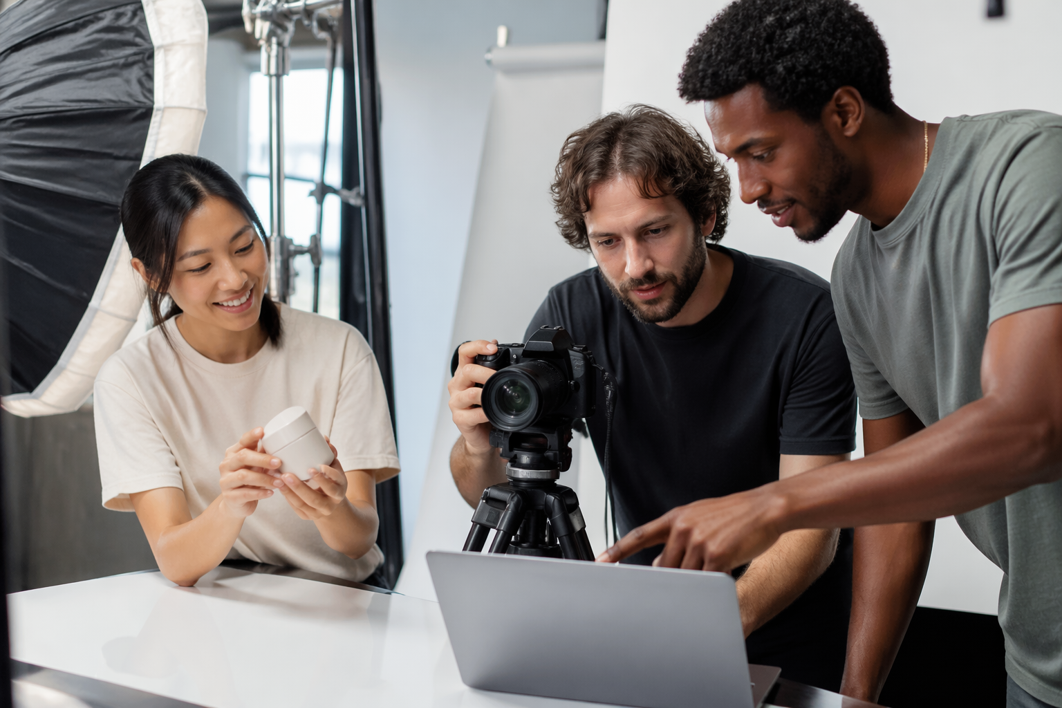 Diverse team of three collaborating on a product video shoot in a bright, modern e-commerce studio with softbox lighting, a tripod-mounted camera, and a laptop showing a video timeline