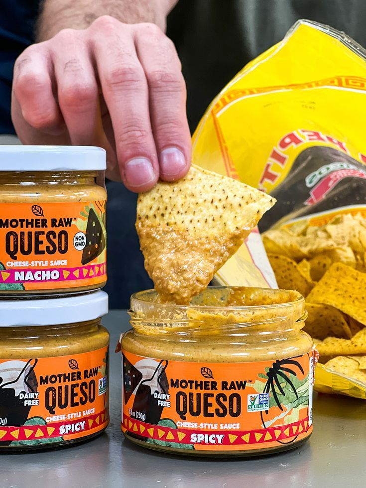 A person dips a tortilla chip into a jar of spicy queso, surrounded by other dip jars and a bag of chips.